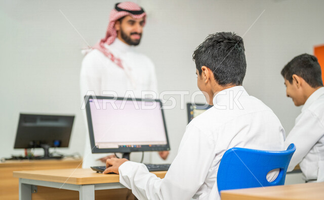 صورة مقربة لمعلم عربي خليجي سعودي في معمل الحاسب الآلي، تعليم الطلبة كيفية استخدام الكمبيوتر في المدرسة، استخدام لوحة المفاتيح للكتابة، التعليم الالكتروني، المناهج المطورة، المشاركة الفعالة للطلاب، التعليم الفعال، التعليم في المملكة العربية السعودية