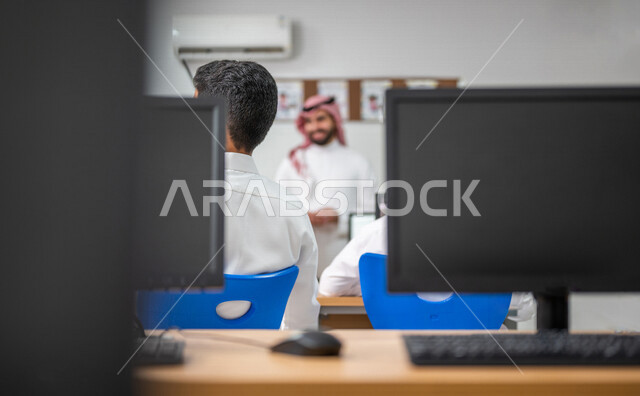 صورة مقربة لشاشات الكمبيوتر، معلم عربي خليجي سعودي في معمل الحاسب الآلي، تعليم الطلبة كيفية استخدام الكمبيوتر في المدرسة، استخدام لوحة المفاتيح للكتابة، التعليم الالكتروني، المناهج المطورة، المشاركة الفعالة للطلاب، التعليم الفعال، التعليم في المملكة العرب