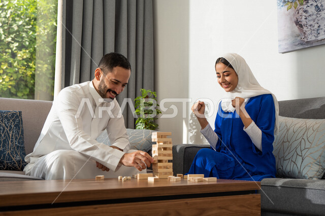 زوجان عربيان خليجيان سعوديان يجلسان في غرفة المعيشة، اللعب في لعبة البناء الخشبيه, لعبة توازن القطع الخشبيه, ألعاب التحدي والتشويق, ممارسة الانشطة الترفيهية، قضاء وقت عائلي ممتع في المنزل، أجواء عائلية سعيدة