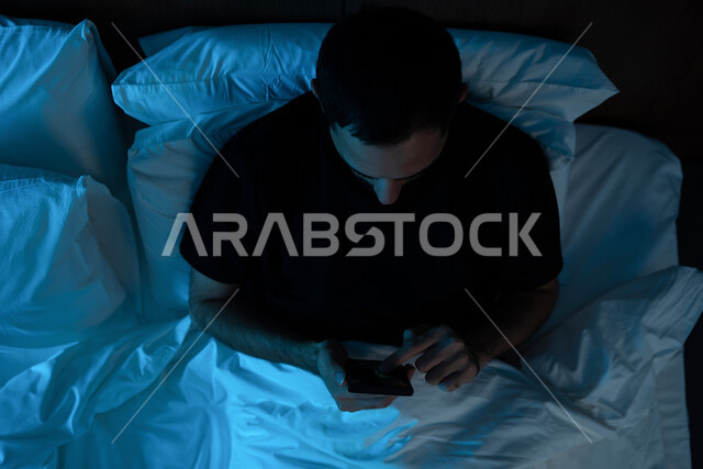 A close-up image of a Saudi Arabian Gulf man lying on his bed, using a mobile phone before bed, surfing the Internet, social networking, completing tasks before bed, using modern technologies