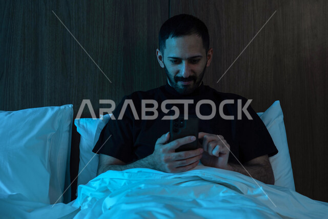 صوره مقربه لرجل عربي خليجي سعودي مستلقي على فراشه،  إستخدام الهاتف المحمول قبل النوم, تصفح الإنترنت, التواصل الإجتماعي, إنجاز المهام قبل النوم، استخدام تقنيات حديثة