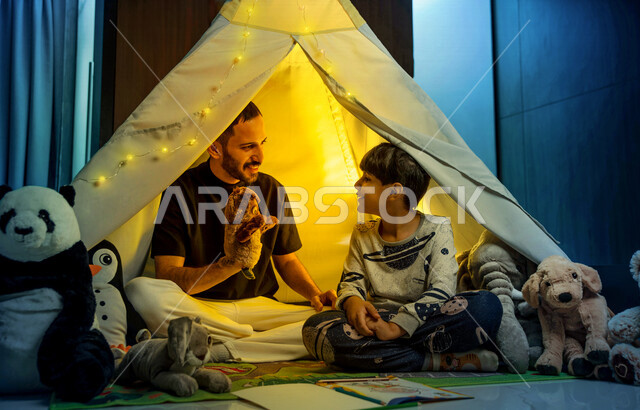 أب مع إبنه عربيان سعوديان خليجيان يجلسان داخل خيمه في وقت الليل في المنزل, الإستمتاع باللعب في المنزل, التخييم في فصل الشتاء, اللعب بالألعاب في غرفة النوم, قضاء وقت ممتع مع العائله, قراءة القصص قبل النوم, أجواء عائليه سعيده