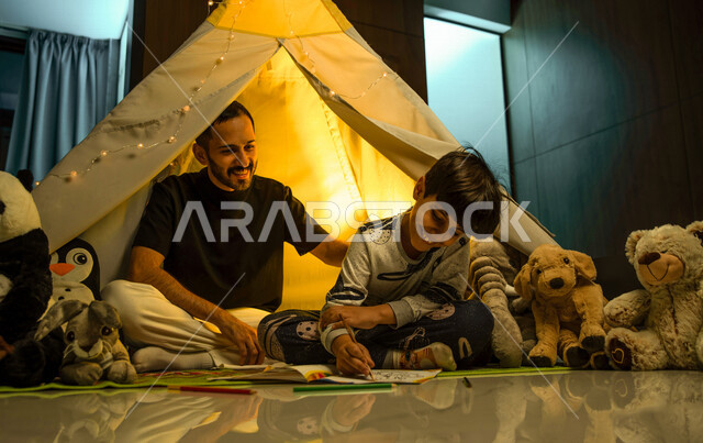أب مع إبنه عربيان سعوديان خليجيان يجلسان داخل خيمه في وقت الليل في المنزل, الإستمتاع باللعب في المنزل, التخييم في فصل الشتاء, اللعب بالألعاب في غرفة النوم, قضاء وقت ممتع مع العائله, قراءة القصص قبل النوم, أجواء عائليه سعيده