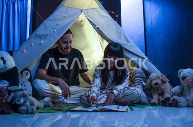 أب مع إبنته عربيان سعوديان خليجيان يجلسان داخل خيمه في وقت الليل في المنزل, الإستمتاع باللعب في المنزل, التخييم في فصل الشتاء, اللعب بالألعاب في غرفة النوم, قضاء وقت ممتع مع العائله, قراءة القصص قبل النوم, أجواء عائليه سعيده