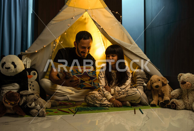أب مع إبنته عربيان سعوديان خليجيان يجلسان داخل خيمه في وقت الليل في المنزل, الإستمتاع باللعب في المنزل, التخييم في فصل الشتاء, اللعب بالألعاب في غرفة النوم, قضاء وقت ممتع مع العائله, قراءة القصص قبل النوم, أجواء عائليه سعيده