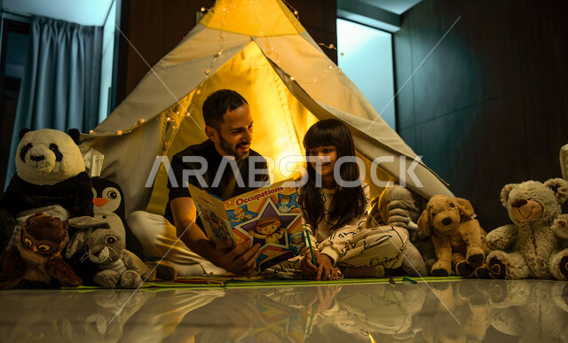 أب مع إبنته عربيان سعوديان خليجيان يجلسان داخل خيمه في وقت الليل في المنزل, الإستمتاع باللعب في المنزل, التخييم في فصل الشتاء, اللعب بالألعاب في غرفة النوم, قضاء وقت ممتع مع العائله, قراءة القصص قبل النوم, أجواء عائليه سعيده