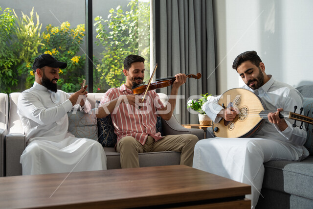 مجموعة من الأصدقاء العرب السعوديين يجلسون في المنزل، العزف على آلة الكمان وآلة العود، توثيق اللحظات عبر الجوال، قضاء أوقات ممتعة مع الأصدقاء، جلسة وناسة وطرب، مواهب فنية، الفنون الموسيقية، آلات موسيقية، أنشطة ترفيهية، زيارات الأهل والأقارب