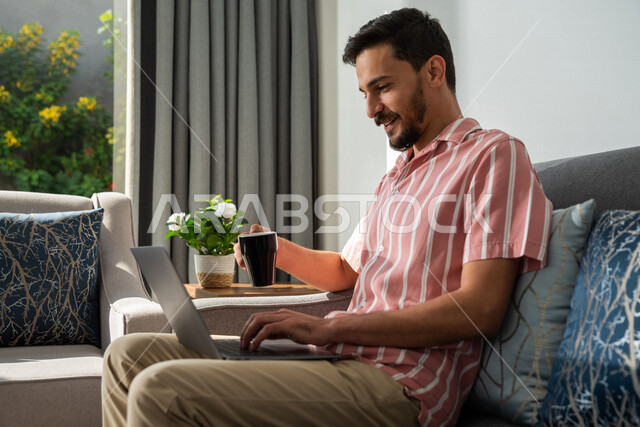 رجل عربي خليجي سعودي يجلس في المنزل،إحتساء القهوة، إستخدام جهاز الحاسوب المحمول، تصفح الإنترنت، جلسة عمل، العمل عن بعد، عقد إجتماع عمل عبر الإنترنت، التسويق الإلكتروني، مهن ووظائف، إجراء مكالمة فيديو، مشاهدة الأفلام والمسلسلات، بث مباشر