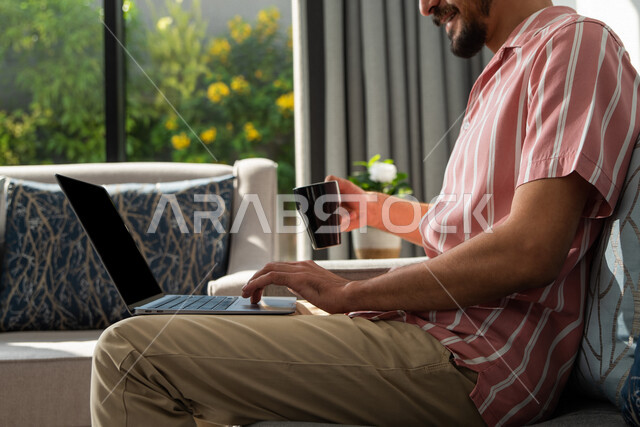 A Saudi Arabian Gulf man sitting at home, drinking coffee, using a laptop, surfing the Internet, working session, working remotely, holding a business meeting online, e-marketing, professions and jobs, making a video call, watching movies and series, live