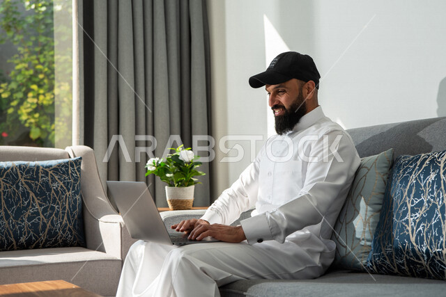 A Saudi Arabian Gulf man sitting at home, using a laptop, browsing the Internet, working session, working remotely, holding a business meeting online, e-marketing, professions and jobs, making a video call, watching movies and series, live broadcasting