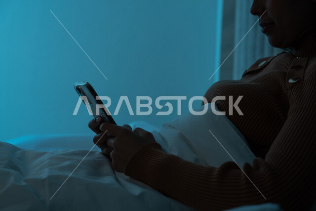 صورة مقربة لامرأة عربية خليجية سعودية ترتدي ملابس الشتاء الدافئة وتستخدم الهاتف المحمول، تصفح الانترنت، التواصل الاجتماعي، مفهوم الراحة والهدوء، الإستمتاع بقضاء أوقات الفراغ، استخدام تقنيات حديثة