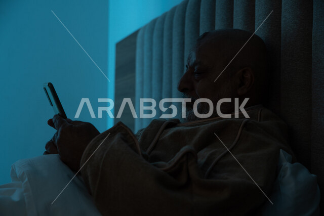 رجل مسن عربي خليجي سعودي يرتدي ملابس الشتاء الدافئة، استخدام الهاتف المحمول، تصفح الانترنت، التواصل الاجتماعي، ارتداء معطف الشتاء، مفهوم الراحة والهدوء، الإستمتاع بقضاء أوقات الفراغ، استخدام تقنيات حديثة