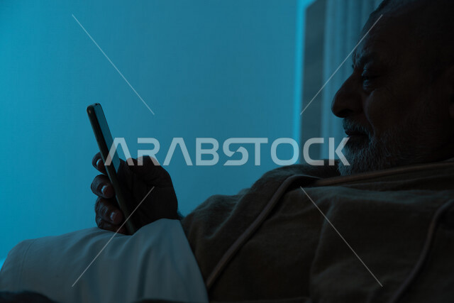 رجل مسن عربي خليجي سعودي يرتدي ملابس الشتاء الدافئة، استخدام الهاتف المحمول، تصفح الانترنت، التواصل الاجتماعي، ارتداء معطف الشتاء، مفهوم الراحة والهدوء، الإستمتاع بقضاء أوقات الفراغ، استخدام تقنيات حديثة