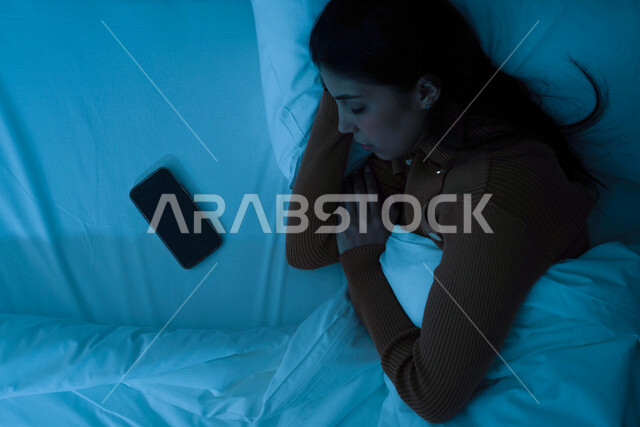 A Saudi Gulf Arab woman wears warm winter clothes while sleeping with a mobile phone with a blank black screen next to her, bedtime, early sleep, a soft white pillow, relaxation and rest, enjoying a quiet and healthy sleep