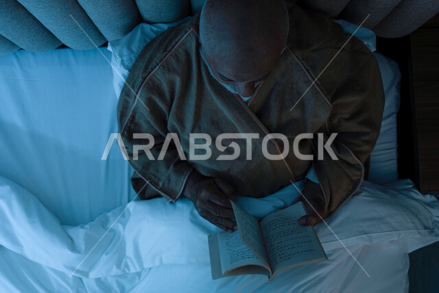 رجل مسن عربي خليجي سعودي يرتدي ملابس الشتاء الدافئة ويحمل بيده كتاب، قراءة الكتب، ارتداء معطف الشتاء، قراءة القصص والروايات، مفهوم الراحة والهدوء، الإستمتاع بقضاء أوقات الفراغ