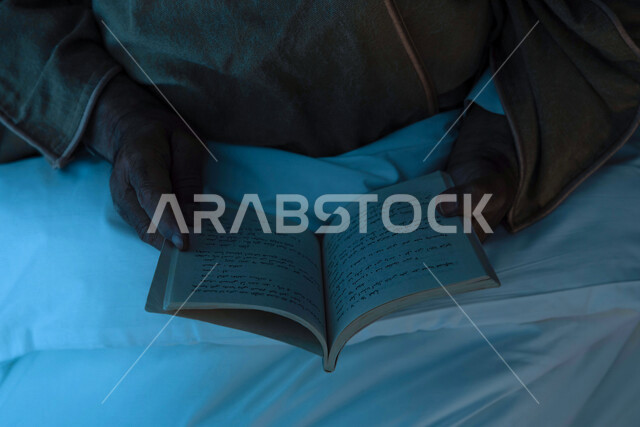 صورة مقربة لرجل مسن عربي خليجي سعودي يرتدي ملابس الشتاء الدافئة ويحمل بيده كتاب، قراءة الكتب، ارتداء معطف الشتاء، قراءة القصص والروايات، مفهوم الراحة والهدوء، الإستمتاع بقضاء أوقات الفراغ