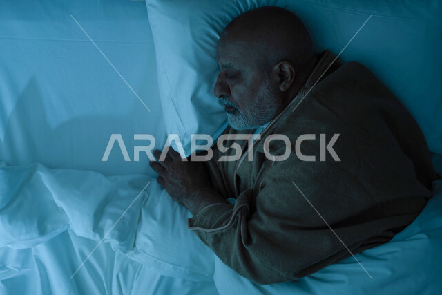 رجل مسن عربي خليجي سعودي يرتدي ملابس الشتاء الدافئة اثناء النوم، وقت النوم، النوم المبكر، ارتداء معطف الشتاء، وسادة بيضاء ناعمة، الاسترخاء والراحة، الاستمتاع بنوم هادئ وصحي