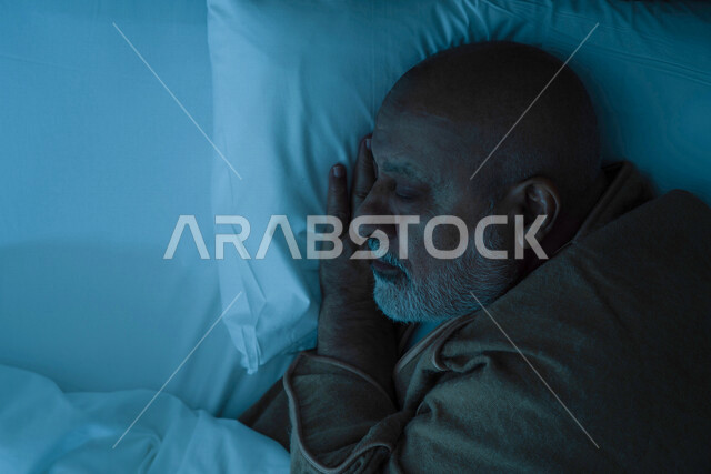 رجل مسن عربي خليجي سعودي يرتدي ملابس الشتاء الدافئة اثناء النوم، وقت النوم، النوم المبكر، ارتداء معطف الشتاء، وسادة بيضاء ناعمة، الاسترخاء والراحة، الاستمتاع بنوم هادئ وصحي