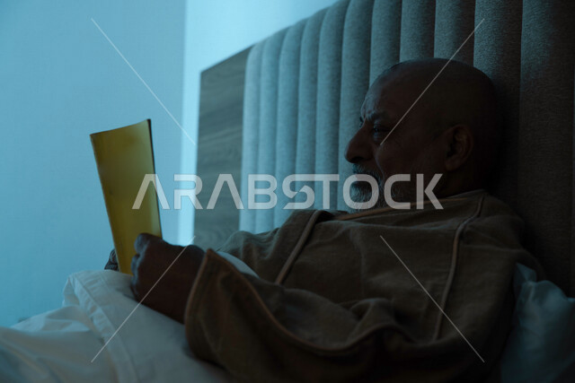 رجل مسن عربي خليجي سعودي يرتدي ملابس الشتاء الدافئة ويحمل بيده كتاب، قراءة الكتب، ارتداء معطف الشتاء، قراءة القصص والروايات، مفهوم الراحة والهدوء، الإستمتاع بقضاء أوقات الفراغ