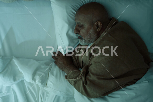 رجل مسن عربي خليجي سعودي يرتدي ملابس الشتاء الدافئة اثناء النوم، وقت النوم، النوم المبكر، ارتداء معطف الشتاء، وسادة بيضاء ناعمة، الاسترخاء والراحة، الاستمتاع بنوم هادئ وصحي