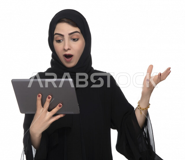 Portrait of an Emirati Arab Gulf woman in traditional dress using a tablet with facial gestures indicating astonishment, e-marketing, the concept of technology and the use of modern technical devices, remote work, video call, watching programs, white back