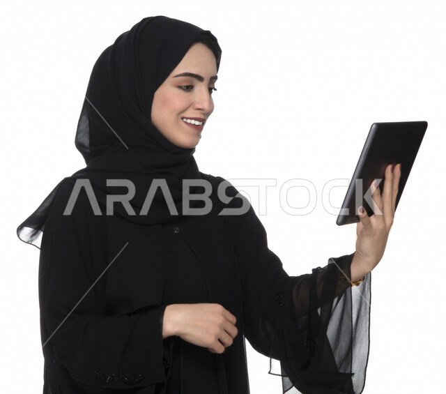 Portrait of an Emirati Arab Gulf woman in traditional dress using a tablet with facial gestures indicating joy, e-marketing, the concept of technology and the use of modern technical devices, remote work, video call, watching programs, white background