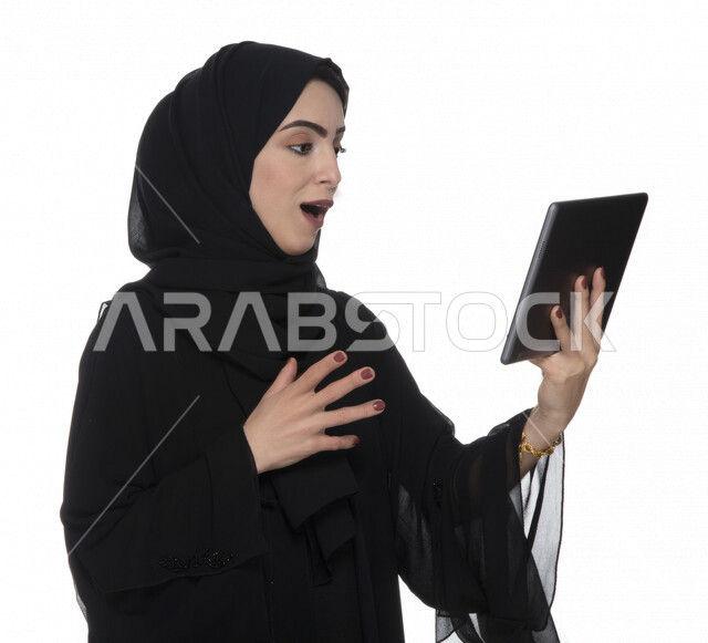 Portrait of an Emirati Arab Gulf woman in traditional dress using a tablet with facial gestures indicating astonishment, e-marketing, the concept of technology and the use of modern technical devices, remote work, video call, watching programs, white back