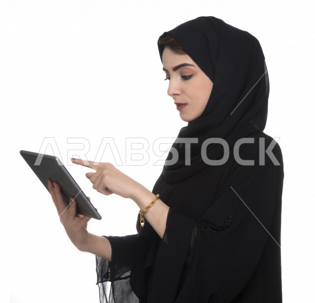 Portrait of an Emirati Arab Gulf woman in traditional dress using a tablet, e-marketing, the concept of technology and the use of modern technical devices, remote work, video call, watching programs, white background