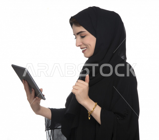 Portrait of an Emirati Arab Gulf woman in traditional dress using a tablet with facial gestures indicating joy, e-marketing, the concept of technology and the use of modern technical devices, remote work, video call, watching programs, white background