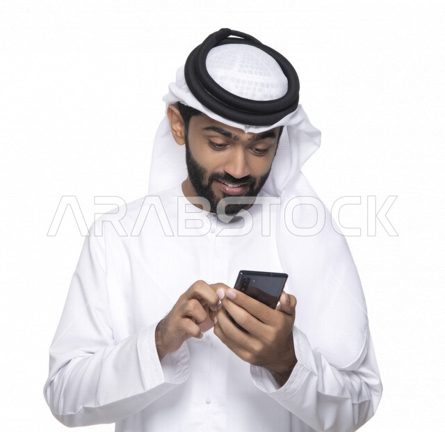 Portrait of an Arab Gulf Emirati youth using a mobile phone, facial gestures indicating joy and happiness, communication with individuals, online orders and marketing, social relations, use of technical devices, white background