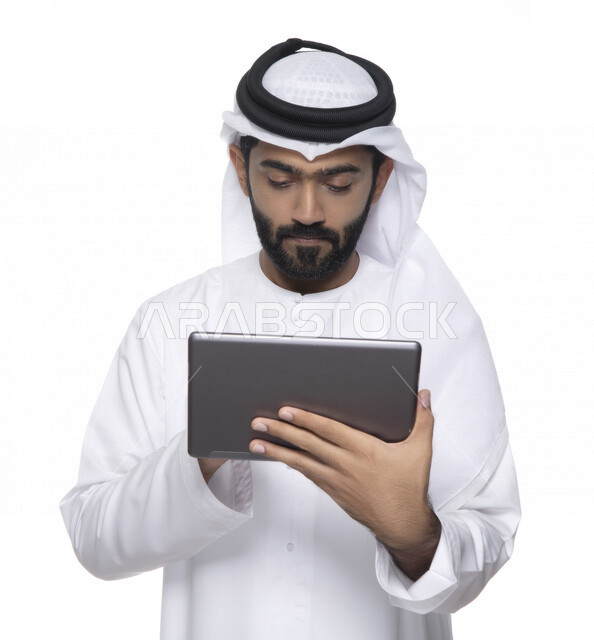 Portrait of an Arab Gulf Emirati man in traditional dress using a tablet, e-marketing, the concept of technology and the use of modern technical devices, remote work, video call, watching programs, white background