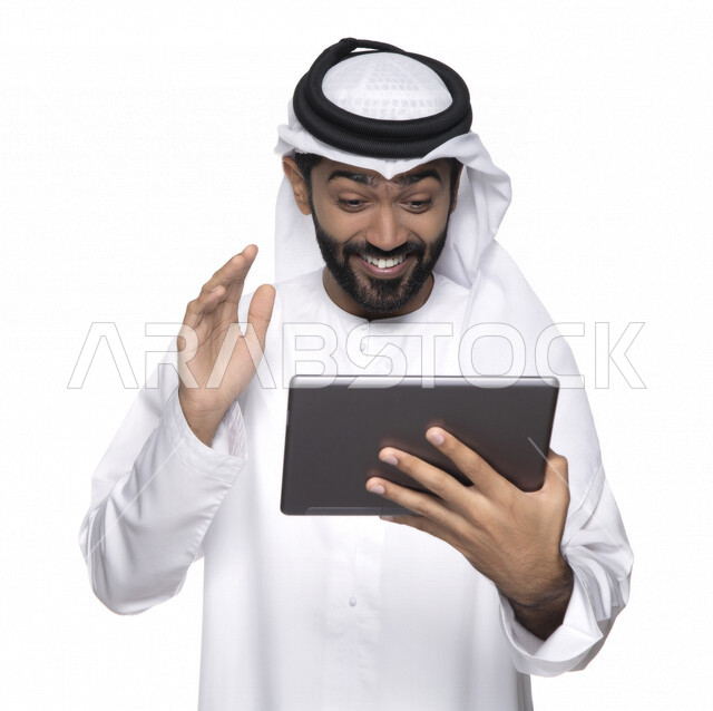 Portrait of an Arab Gulf Emirati man in traditional dress using a tablet, e-marketing, the concept of technology and the use of modern technical devices, remote work, video call, watching programs, white background