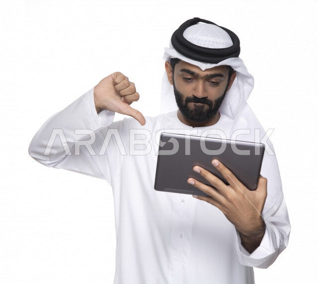 Portrait of an Arab Gulf Emirati man in traditional attire using a tablet and pointing his thumb downwards, gestures indicating dislike, the concept of technology and the use of modern technical devices, working remotely, watching programs, a white backgr