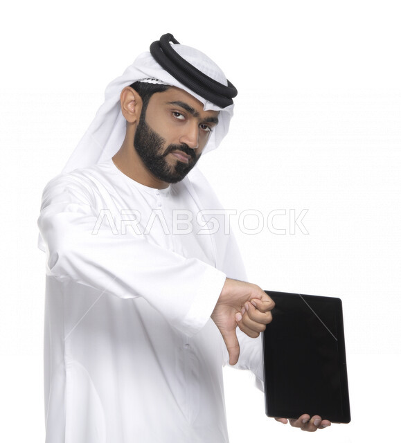 Portrait of an Arab Gulf Emirati man in traditional attire using a tablet and pointing his thumb downwards, gestures indicating dislike, the concept of technology and the use of modern technical devices, working remotely, watching programs, a white backgr