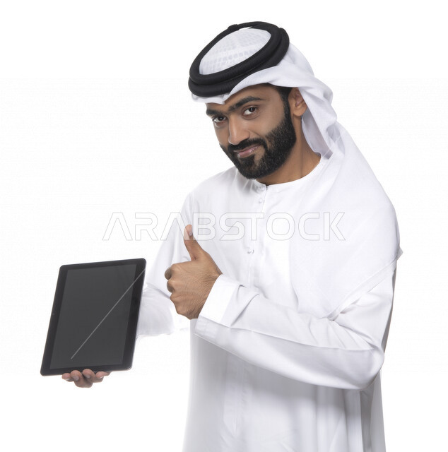 Portrait of an Arab Gulf Emirati man in traditional attire using a tablet and pointing his thumb up, gestures indicating admiration, e-marketing, remote work, video call, watching programmes, white background