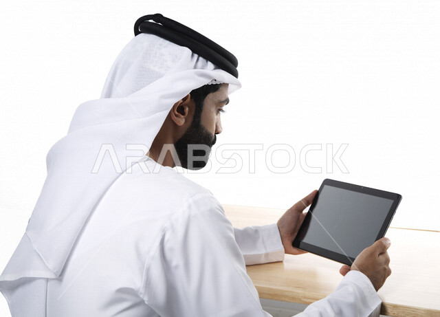 Portrait of an Arab Gulf Emirati businessman, using a tablet with a flat black screen, completing work tasks, surfing the Internet, working session, holding a business meeting via the Internet, e-marketing, white background