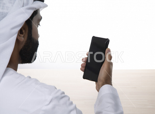 A close-up of an Arab Gulf Emirati man using a mobile phone, online shopping, surfing the Internet, remote video call, using modern technology devices, a white background