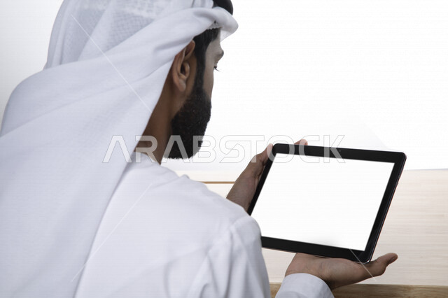Portrait of an Arab Gulf Emirati businessman, using a tablet with a white flat screen, completing work tasks, surfing the Internet, working session, holding a business meeting via the Internet, e-marketing, white background