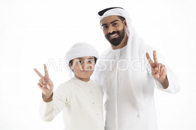 بورتريه لرجل عربي إماراتي خليجي يرتدي الزي التقليدي بجانب طفله , يأشران بيدهم علامة النصر ، تجمع عائلي ، تعابير وجه مبتسمة  ، قضاء أوقات عائلية جميلة  ، خلفية بيضاء