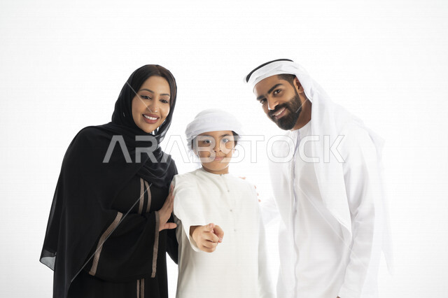 بورتريه لزوجان عربيان خليجيان إماراتيان بجانب إبنهم يرتديان  الزي الإماراتي التقليدي , قضاء وقت ممتع مع العائلة , عائلة مبتسمة , طفل إماراتي بعمامة وكندورة  يأشر بيده أمام الكاميرا  ,  خلفية بيضاء 