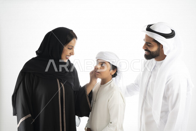 بورتريه لزوجان عربيان خليجيان إماراتيان بجانب إبنهم يرتديان  الزي الإماراتي التقليدي , قضاء وقت ممتع مع العائلة , إيماءات وجه تدل على السعادة والفرح , حضن الإبن , أم تمسك بخد ولدها بكل حب وحنان  , خلفية بيضاء 