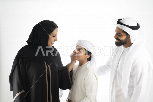 بورتريه لزوجان عربيان خليجيان إماراتيان بجانب إبنهم يرتديان  الزي الإماراتي التقليدي , قضاء وقت ممتع مع العائلة , إيماءات وجه تدل على السعادة والفرح , حضن الإبن , أم تمسك بخد ولدها بكل حب وحنان  , خلفية بيضاء 