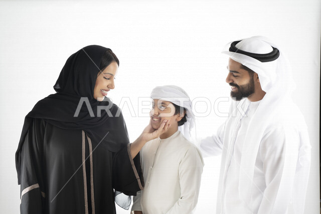 بورتريه لزوجان عربيان خليجيان إماراتيان بجانب إبنهم يرتديان  الزي الإماراتي التقليدي , قضاء وقت ممتع مع العائلة , إيماءات وجه تدل على السعادة والفرح , حضن الإبن , أم تمسك بخد ولدها بكل حب وحنان  , خلفية بيضاء 