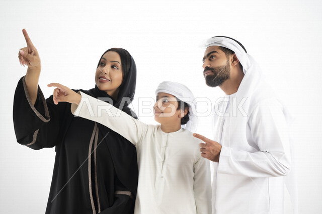 بورتريه لزوجان عربيان خليجيان إماراتيان بجانب إبنهم يرتديان  الزي الإماراتي التقليدي , قضاء وقت ممتع مع العائلة , إيماءات وجه تدل على السعادة والفرح ,يأشران بيدهم إلى الأعلى ,  خلفية بيضاء 