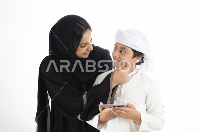بورتريه لأم عربية خليجية إماراتية ترتدي الزي التقليدي الإماراتي شيلة وعباية ، تعليم الطفل كيفية استخدام هاتف المحمول ، استخدام خاصية اللمس لاستعمال الجهاز ، التعليم الالكتروني، التعليم الفعال، خلفية بيضاء 