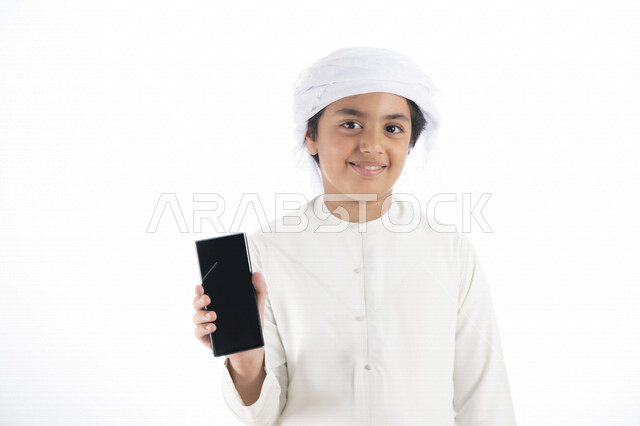 بورتريه لطفل عربي إماراتي خليجي يرتدي كندورة وعمامة إماراتيه , يحمل بيده هاتف المحمول بشاشة سوداء مفرغة   ، التسلية والترفيه ، تطبيقات الهاتف الخلفي ,  خلفية بيضاء