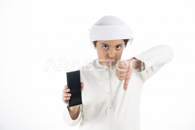 Portrait of an Arab Emirati Gulf child wearing an Emirati kandura and turban, holding a mobile phone with a hollow black screen in his hand, pointing his thumb downwards, facial gestures indicating usurpation, back-phone applications, white background