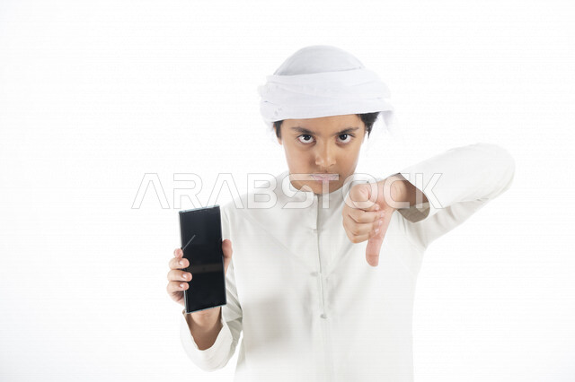 بورتريه لطفل عربي إماراتي خليجي يرتدي كندورة وعمامة إماراتيه , يحمل بيده هاتف المحمول بشاشة سوداء مفرغة   ، يأشر بأصبع الإبهام للأسفل , إيماءات وجه تدل على الغصب  ، تطبيقات الهاتف الخلفي ,  خلفية بيضاء