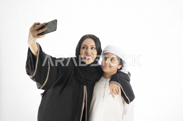 بورتريه لأم وإبن عربي خليجي إماراتي يرتديان الزي التقليدي ,  مستمتعتان بالتقاط صورة سيلفي من خلال الهاتف المحمول، التقاط صور تذكارية ، صورة جماعية ، توثيق اللحظات السعيدة ، خلفية بيضاء 