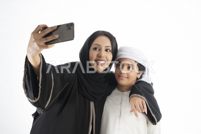 بورتريه لأم وإبن عربي خليجي إماراتي يرتديان الزي التقليدي ,  مستمتعتان بالتقاط صورة سيلفي من خلال الهاتف المحمول، التقاط صور تذكارية ، صورة جماعية ، توثيق اللحظات السعيدة ، خلفية بيضاء 